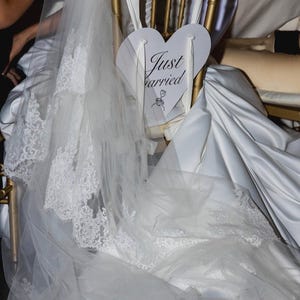 May include: A gold-toned chair features a white heart-shaped sign with the text Just Married and a small rose illustration. A long white wedding veil with intricate lace trim drapes over the back of the chair, partially covering a white satin fabric.