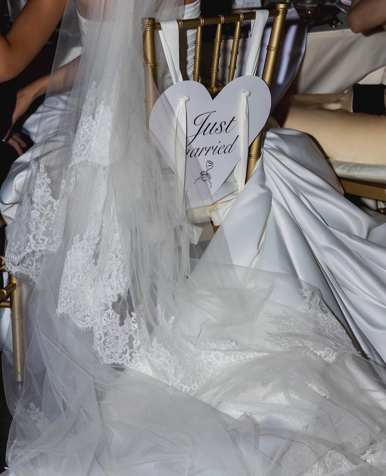 May include: A gold-toned chair features a white heart-shaped sign with the text Just Married and a small rose illustration. A long white wedding veil with intricate lace trim drapes over the back of the chair, partially covering a white satin fabric.