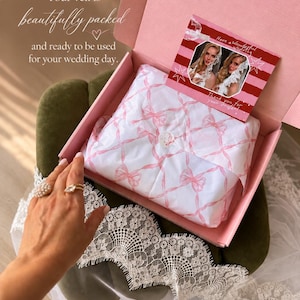 May include: A pink cardboard box containing white tissue paper with a pink ribbon pattern, sealed with a circular sticker. A rectangular card with red stripes and two photos of a person wearing a white lace veil rests inside the box.