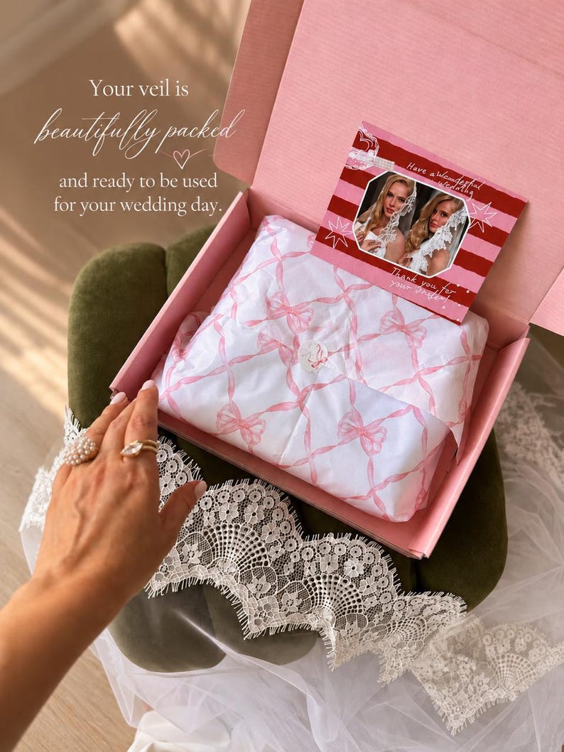 May include: A pink cardboard box containing white tissue paper with a pink ribbon pattern, sealed with a circular sticker. A rectangular card with red stripes and two photos of a person wearing a white lace veil rests inside the box.