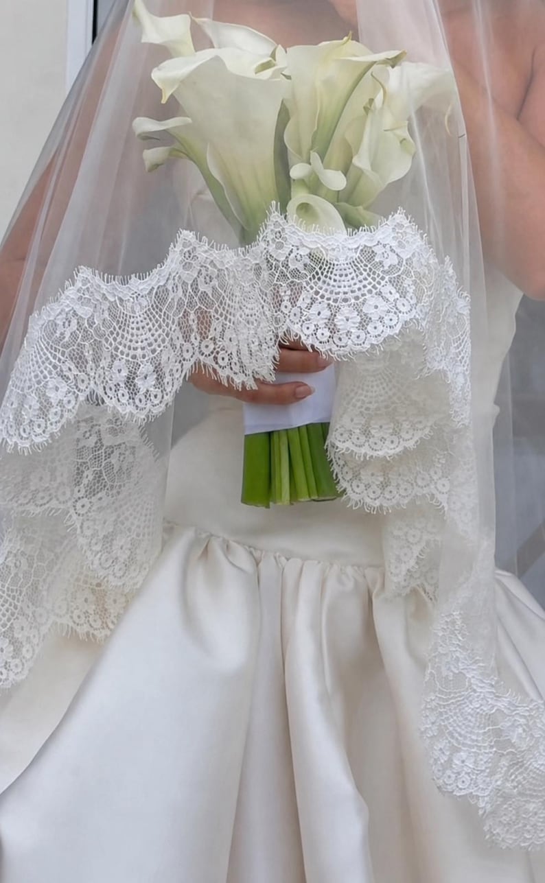 May include: A white wedding veil featuring an intricate scalloped lace border with floral patterns, draped over a bouquet of white calla lilies with green stems wrapped in white ribbon, worn with a smooth, solid white satin dress.