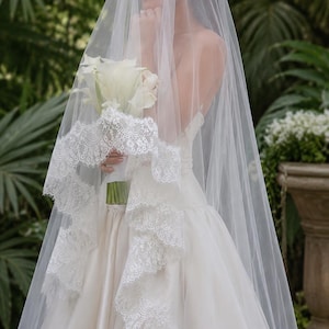 May include: A long, sheer white wedding veil featuring a wide, intricate floral lace trim along the edges. The veil is draped over a white satin gown, and a bouquet of white calla lilies is held in the center, partially covered by the tulle fabric.