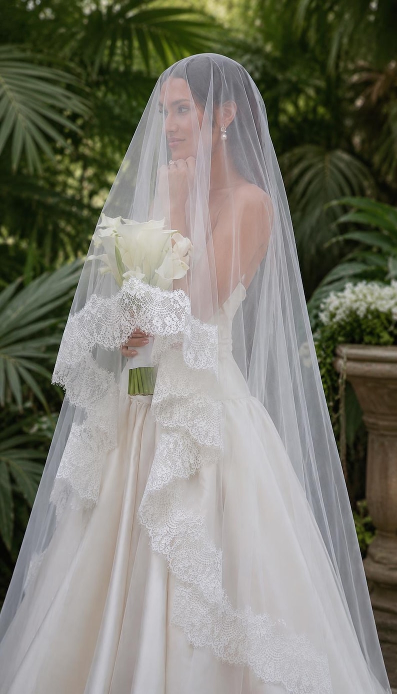 May include: A long, sheer white wedding veil featuring a wide, intricate floral lace trim along the edges. The veil is draped over a white satin gown, and a bouquet of white calla lilies is held in the center, partially covered by the tulle fabric.