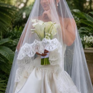 May include: A sheer white bridal veil featuring an intricate scalloped lace trim along the edges. The veil is draped over a strapless ivory satin gown, paired with a bouquet of white calla lilies with green stems, creating a classic ensemble.