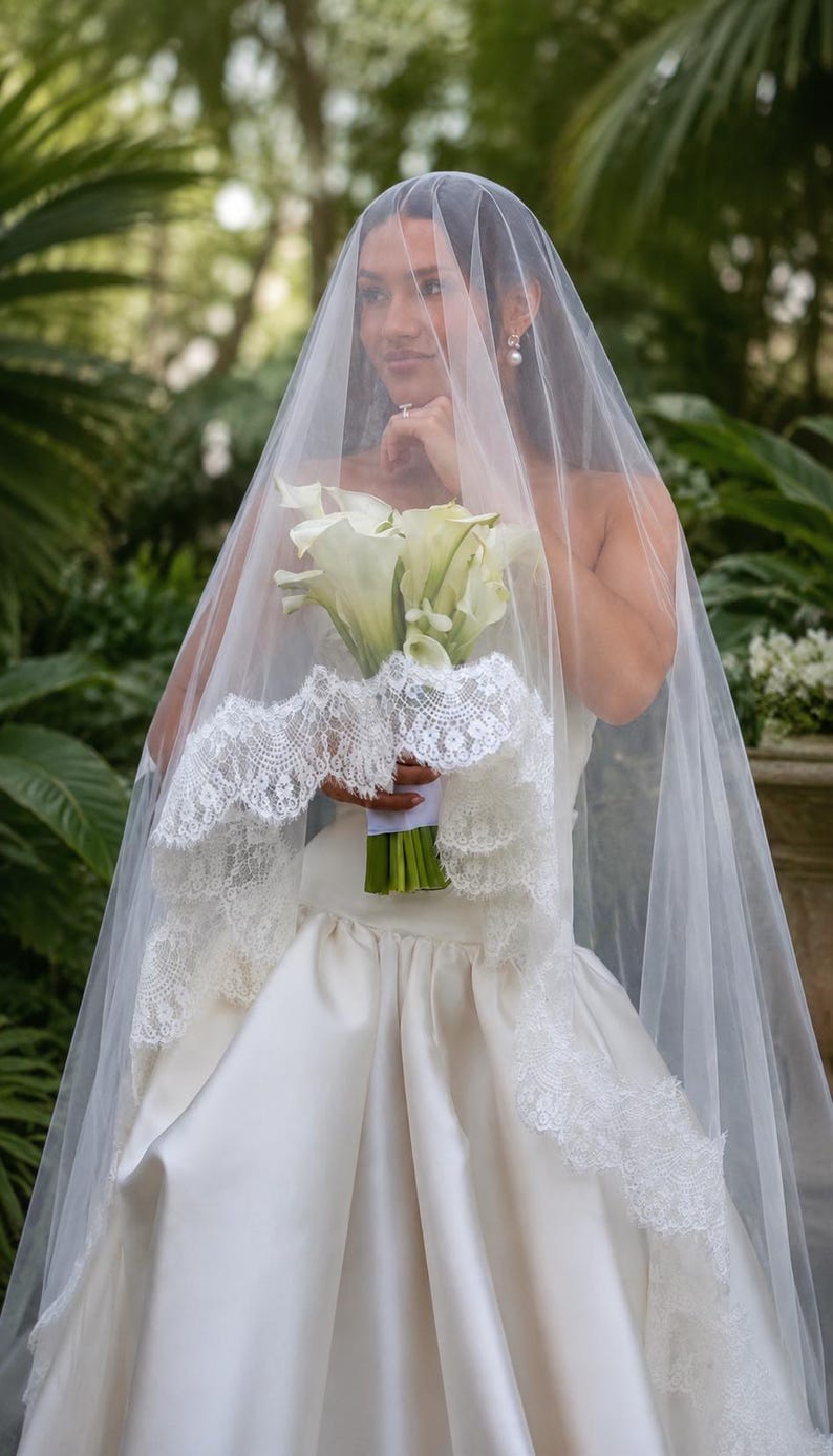 May include: A sheer white bridal veil featuring an intricate scalloped lace trim along the edges. The veil is draped over a strapless ivory satin gown, paired with a bouquet of white calla lilies with green stems, creating a classic ensemble.