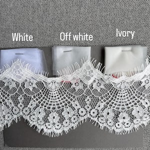 May include: A wide strip of white lace trim with a scalloped edge featuring floral patterns, positioned below three fabric swatches labeled White, Off white, and Ivory. Each swatch is secured with a pin, showing subtle color variations in the tulle.