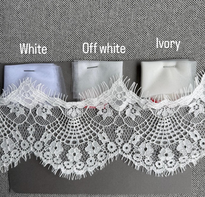 May include: A wide strip of white lace trim with a scalloped edge featuring floral patterns, positioned below three fabric swatches labeled White, Off white, and Ivory. Each swatch is secured with a pin, showing subtle color variations in the tulle.