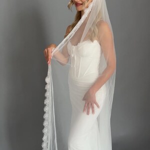 May include: A white bridal gown featuring a corset bodice and a flowing skirt. The bride is wearing a sheer veil with a delicate lace trim. The veil drapes down the back, creating an elegant and romantic look. The dress is a classic bridal style.