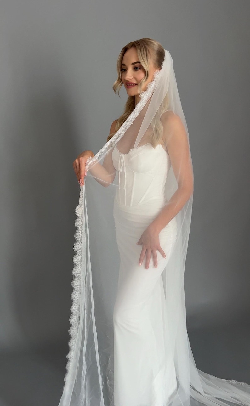 May include: A white bridal gown featuring a corset bodice and a flowing skirt. The bride is wearing a sheer veil with a delicate lace trim. The veil drapes down the back, creating an elegant and romantic look. The dress is a classic bridal style.