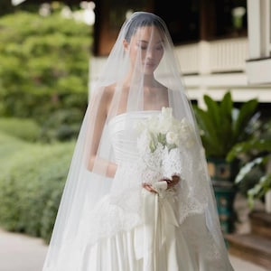 May include: A strapless white satin gown with a full skirt, paired with a long, sheer tulle veil that drapes over the head. The ensemble includes a bouquet of white tulips wrapped in white lace fabric and tied with a long, matching satin ribbon.