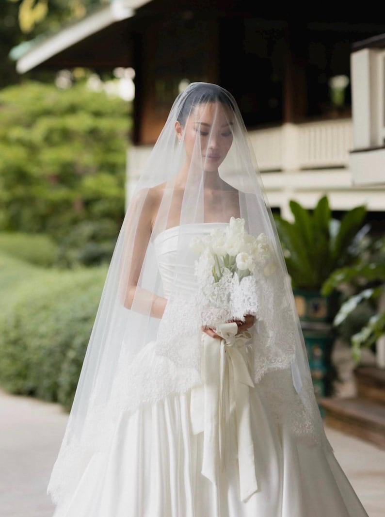 May include: A strapless white satin gown with a full skirt, paired with a long, sheer tulle veil that drapes over the head. The ensemble includes a bouquet of white tulips wrapped in white lace fabric and tied with a long, matching satin ribbon.