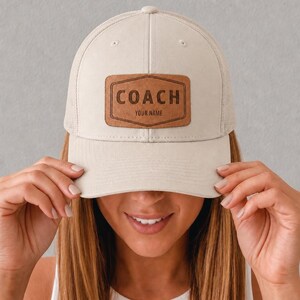 Può includere: Cappellino da camionista color crema con retro in rete e visiera curva. La parte anteriore presenta una toppa in pelle marrone con la scritta "COACH" e il testo "YOUR NAME" in un carattere serif. Il cappello è tenuto da una persona.