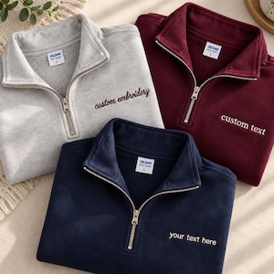 May include: Three Gildan quarter-zip sweatshirts in light gray, maroon, and navy blue. Each features a metal zipper, a collar, and embroidered text on the chest reading custom embroidery, custom text, and your text here respectively.