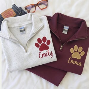 Puede incluir: Dos sudaderas con cremallera de cuarto, una gris claro y otra granate, cada una con una huella bordada y un nombre. La sudadera gris tiene el nombre Emily en granate, y la sudadera granate tiene el nombre Emma en color dorado.