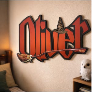 May include: A wooden wall hanging featuring the name "Oliver" in large, red, stylized letters with a black outline. A brown witch's hat and a broom are attached to the sign. The broom is brown with gold accents. The sign is mounted on a beige wall.