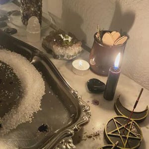 May include: A dimly lit scene featuring a silver tray with salt, lit candles, crystals, and a small jar with wooden shapes. A black candle burns near a triple moon symbol and a pentagram.
