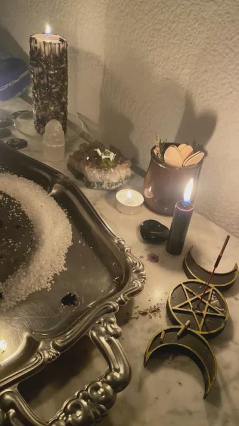 May include: A dimly lit scene featuring a silver tray with salt, lit candles, crystals, and a small jar with wooden shapes. A black candle burns near a triple moon symbol and a pentagram.