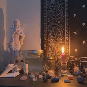 May include: A dimly lit scene featuring a white statue, glass jars, crystals, and a burning candle. A black tapestry with sun and star designs hangs in the background. The items are arranged on a dark surface.