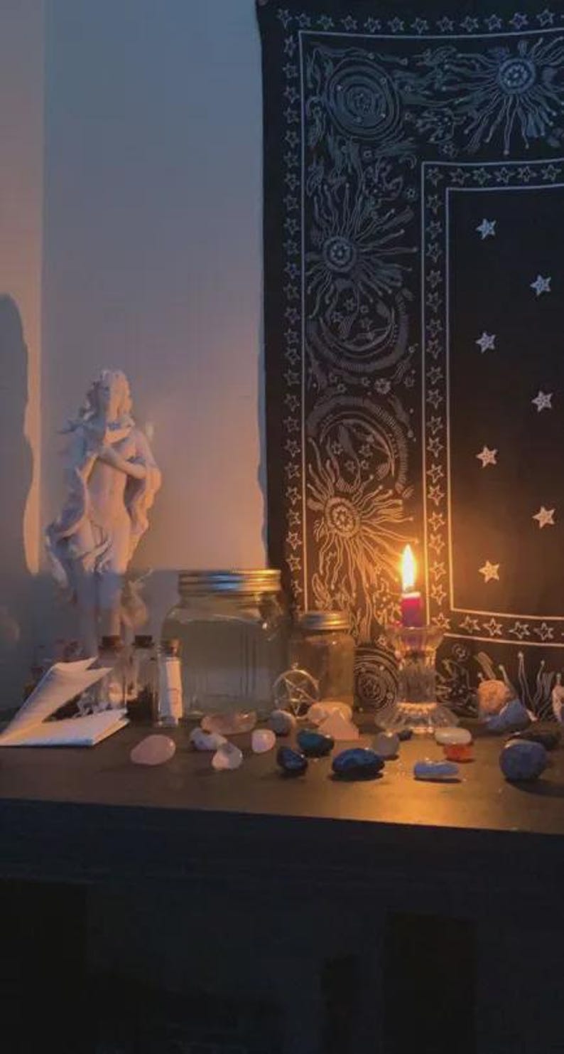 May include: A dimly lit scene featuring a white statue, glass jars, crystals, and a burning candle. A black tapestry with sun and star designs hangs in the background. The items are arranged on a dark surface.