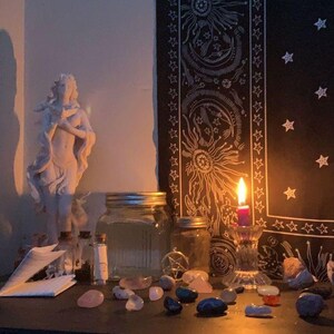 Puede incluir: Una escena tenue con una estatua blanca, frascos de vidrio, cristales y una vela encendida. Un tapiz negro con dise&ntilde;os de sol y estrellas cuelga en el fondo. Los objetos est&aacute;n dispuestos sobre una superficie oscura.