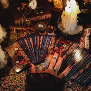 May include: A hand with long red nails wearing multiple rings rests on a deck of tarot cards. The cards have ornate designs with a bird and eye motif. A lit candle and antique items are in the background.