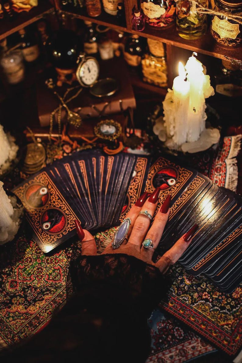 May include: A hand with long red nails wearing multiple rings rests on a deck of tarot cards. The cards have ornate designs with a bird and eye motif. A lit candle and antique items are in the background.