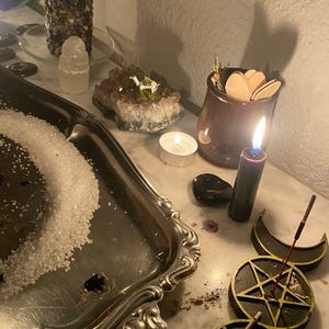 以下が含まれることがあります： 点灯した黒いろうそく、塩の入った銀色のトレイ、装飾的な五芒星が特徴の神秘的な配置。 他の要素には、小さな点灯したろうそく、クリスタル、木製の形が入った瓶が含まれており、スピリチュアルな雰囲気を醸し出しています。