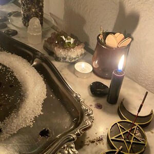 Puede incluir: Una disposici&oacute;n m&iacute;stica con una vela negra encendida, una bandeja plateada con sal y un pentagrama decorativo. Otros elementos incluyen una peque&ntilde;a vela encendida, cristales y un tarro con formas de madera, creando un ambiente espiritual.