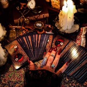 Puede incluir: Una mano con u&ntilde;as largas rojas, adornada con anillos, descansa sobre una baraja de cartas del tarot. Las cartas tienen dise&ntilde;os ornamentados con detalles en rojo y dorado. Velas encendidas y objetos antiguos crean una atm&oacute;sfera m&iacute;stica.