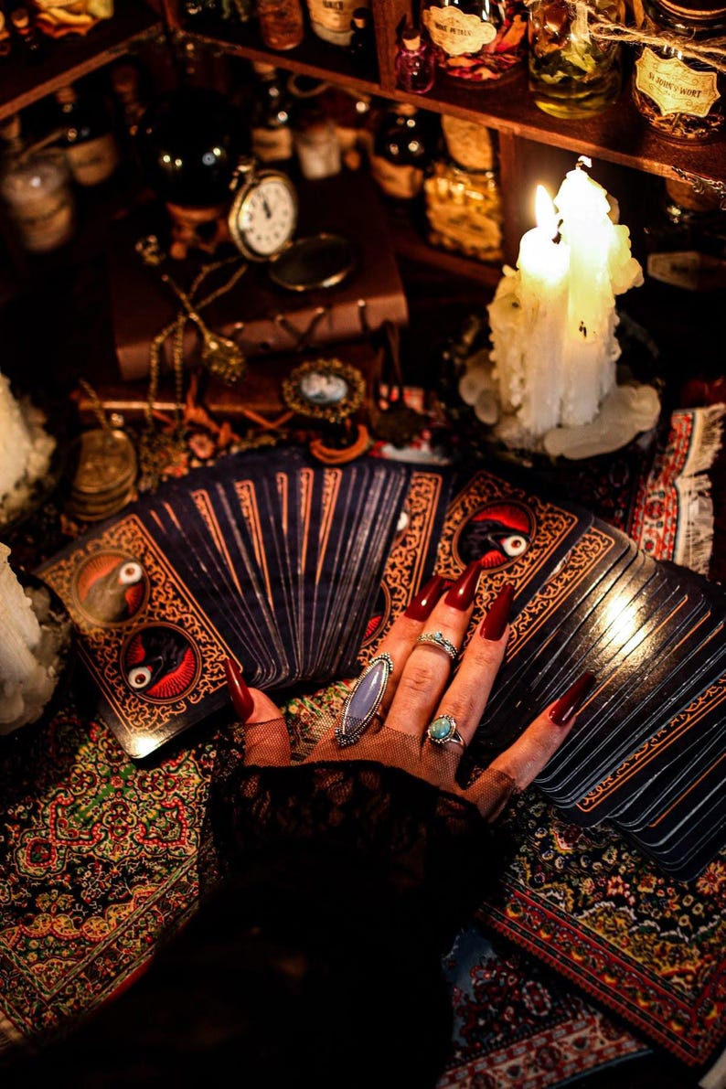 Puede incluir: Una mano con u&ntilde;as largas rojas, adornada con anillos, descansa sobre una baraja de cartas del tarot. Las cartas tienen dise&ntilde;os ornamentados con detalles en rojo y dorado. Velas encendidas y objetos antiguos crean una atm&oacute;sfera m&iacute;stica.