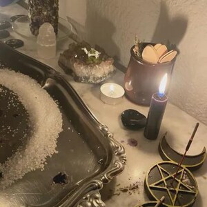 May include: A dimly lit scene featuring a silver tray with salt, lit candles, crystals, and a small jar with wooden shapes. A black candle burns near a triple moon symbol and a pentagram.
