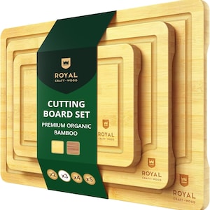 May include: A set of four bamboo cutting boards in varying sizes, nested together. The boards have a light wood grain finish and a recessed juice groove. A dark green label reads "CUTTING BOARD SET" and "PREMIUM ORGANIC BAMBOO". The brand name "ROYAL CRAFT-WOOD" is also visible.