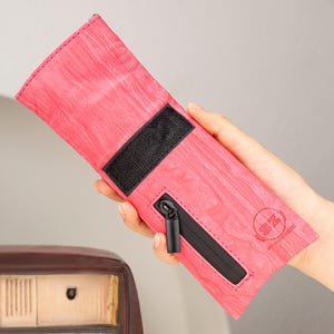 May include: A pink, rectangular case with a black zipper and a black velcro closure. The case has a wood grain texture and a small black zipper pull. The case is held in a hand.