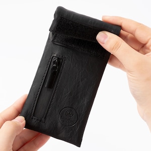 Puede incluir: Una funda de cuero negro con cierre de cremallera y correa de velcro. La funda está sujeta por dos manos sobre un fondo blanco. La funda tiene una superficie texturizada y un pequeño logotipo en la parte inferior.