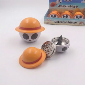 May include: A novelty grinder with a white base, black eye-shaped details, and an orange hat with a red band. The grinder is disassembled, revealing a metal grinding component and a small black scoop. The packaging reads "Stasheeze Grinder."