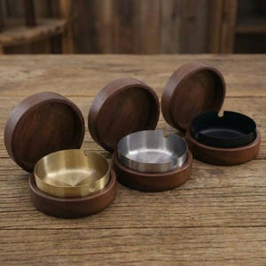 May include: A collection of wooden ashtrays with metal inserts. The ashtrays have a dark brown wooden base and lid, with metal inserts in gold, silver, and black. The ashtrays are arranged on a wooden surface.