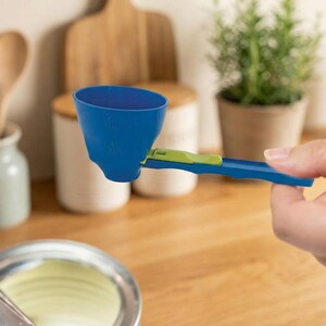 May include: A blue and green adjustable measuring scoop. The scoop is held by a hand and is positioned over a bowl. The scoop has measurements printed on the side. The handle is blue with a green slider.