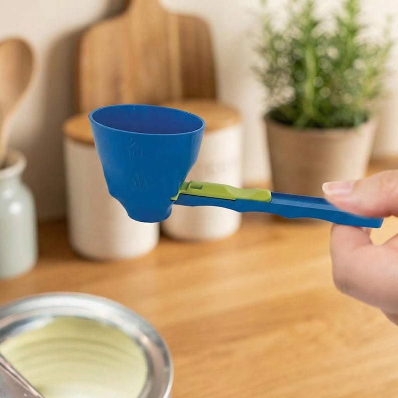 May include: A blue and green adjustable measuring scoop. The scoop is held by a hand and is positioned over a bowl. The scoop has measurements printed on the side. The handle is blue with a green slider.
