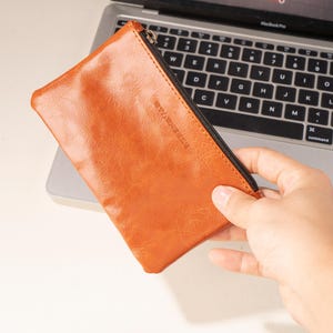 以下が含まれることがあります： 黒いジッパーが付いた長方形のオレンジブラウンの革製ポーチ。ラップトップのキーボードの上に持たれています。ポーチは微妙な質感で、小さなエンボス加工のロゴが付いています。ラップトップの画面にはビデオインターフェースが表示されています。
