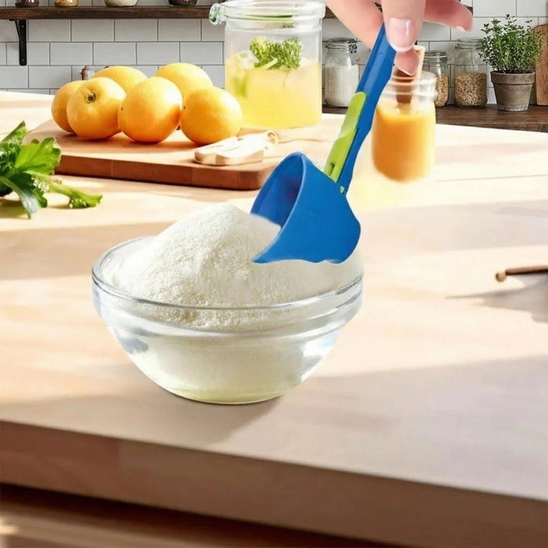 May include: A blue and green measuring scoop is filling a clear glass bowl with white powder. In the background, there are lemons and a wooden chopping board, indicating a kitchen setting.