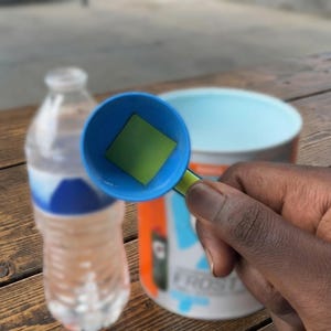 May include: A blue and green measuring scoop holding a green square. A clear water bottle and a container with orange, blue, and white accents are in the background. The scoop is held by a person.