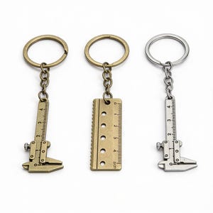 May include: Three metal keyrings with ruler and caliper designs. One is silver, two are bronze-toned. Each keyring features a ring and chain. The ruler keyring has a 5 cm scale. The caliper keyrings have a 4 cm scale.