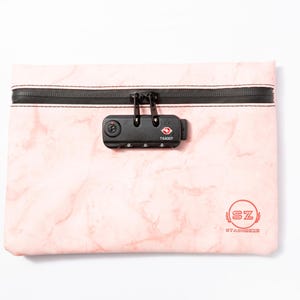 Puede incluir: Una bolsa rosa con estampado de mármol, cremallera negra y cerradura de combinación negra. La cerradura tiene el texto "TSA007". La bolsa tiene un logotipo circular rojo con el texto "SZ STASHED".