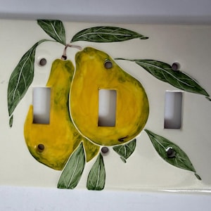 Pode incluir: Placa de interruptor de cerâmica com um desenho pintado à mão de duas peras amarelas com folhas verdes. A placa tem três aberturas para interruptores e é montada sobre um fundo creme. As peras têm sombreamento e destaques detalhados.