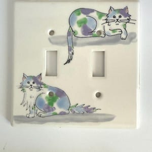 May include: A white ceramic double light switch cover featuring two whimsical cat illustrations. The cats are painted with blue, green, and purple spots. The cover has two switch openings and two screw holes.