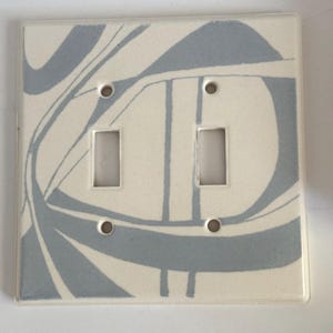 May include: A ceramic double light switch cover with a cream background and a blue abstract design. The cover has two rectangular switch openings and four screw holes for mounting.