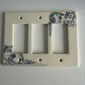 May include: A cream-colored triple light switch cover with hand-painted cat designs. The cats have blue, green, and purple markings. The switch plate has three rectangular openings and four screw holes.