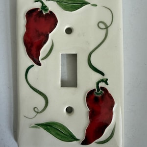 May include: A rectangular off-white light switch plate featuring a hand-painted design of two dark red chilli peppers, green leaves, and thin green vines. The plate has a central rectangular cutout for a toggle switch and two circular screw holes.