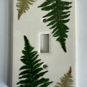 Pode incluir: Uma placa retangular de interruptor de luz em tom branco sujo com um design botânico pintado à mão de quatro folhas de samambaia em vários tons de verde e castanho. A placa inclui um recorte central retangular e dois orifícios para parafusos.