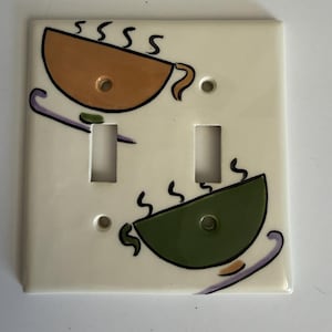 Könnte beinhalten: Eine weiße Doppel-Lichtschalterabdeckung mit einem handgemalten Motiv von zwei Kaffeetassen. Eine braune Tasse befindet sich oben links, eine grüne Tasse unten rechts. Über beiden Tassen ist jeweils eine stilisierte Dampfwolke zu sehen.